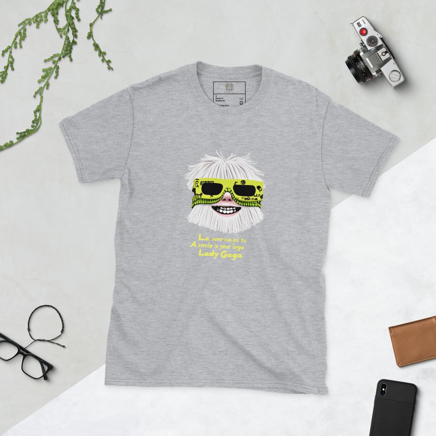 Light grey unisex T-shirt (flat lay) — front print: stylized face with white hair and neon sunglasses, text La sonrisa es tu / A smile is your logo. Lady Gaga.