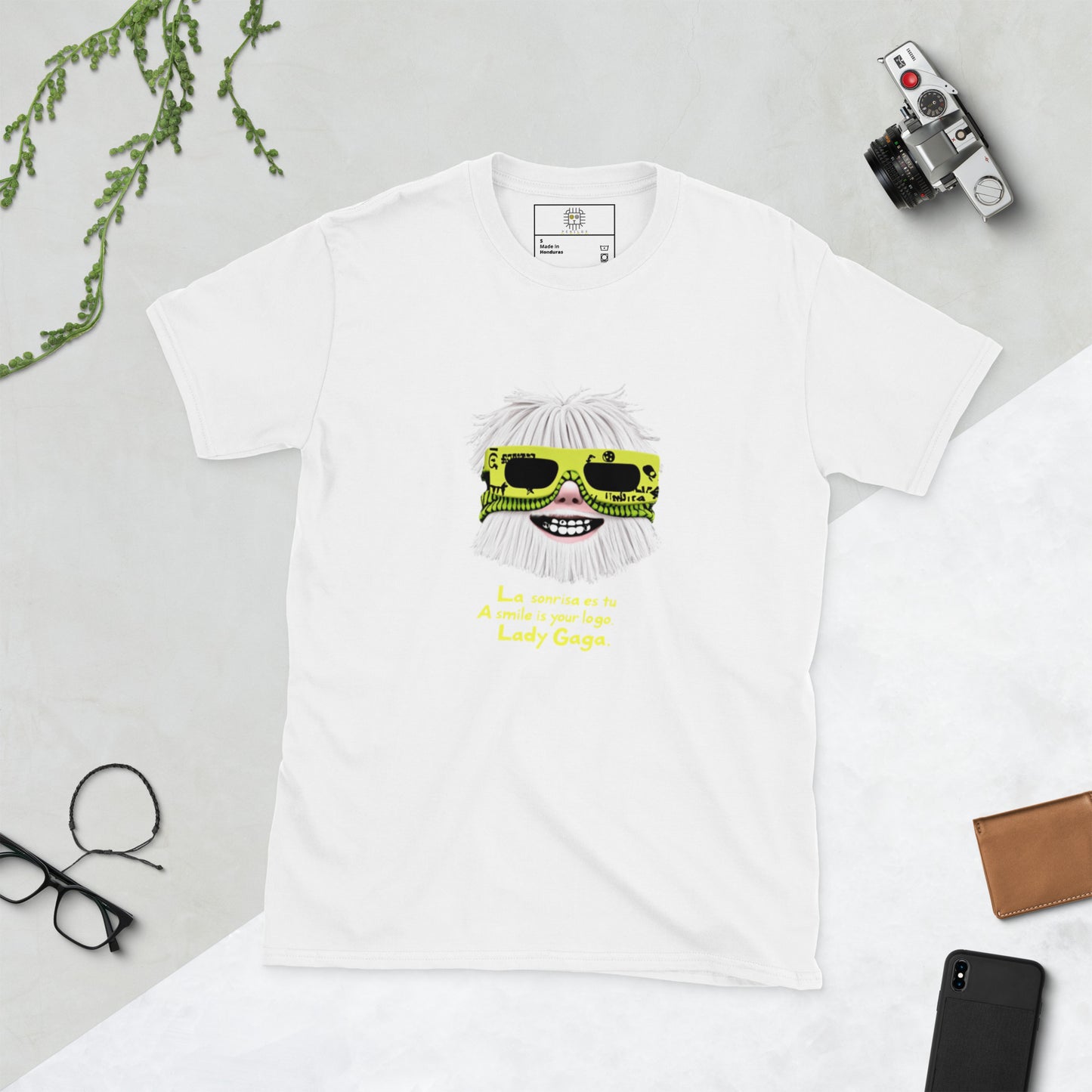 White unisex T-shirt (flat lay) — front print: stylized face with white hair and neon sunglasses, text La sonrisa es tu / A smile is your logo. Lady Gaga.