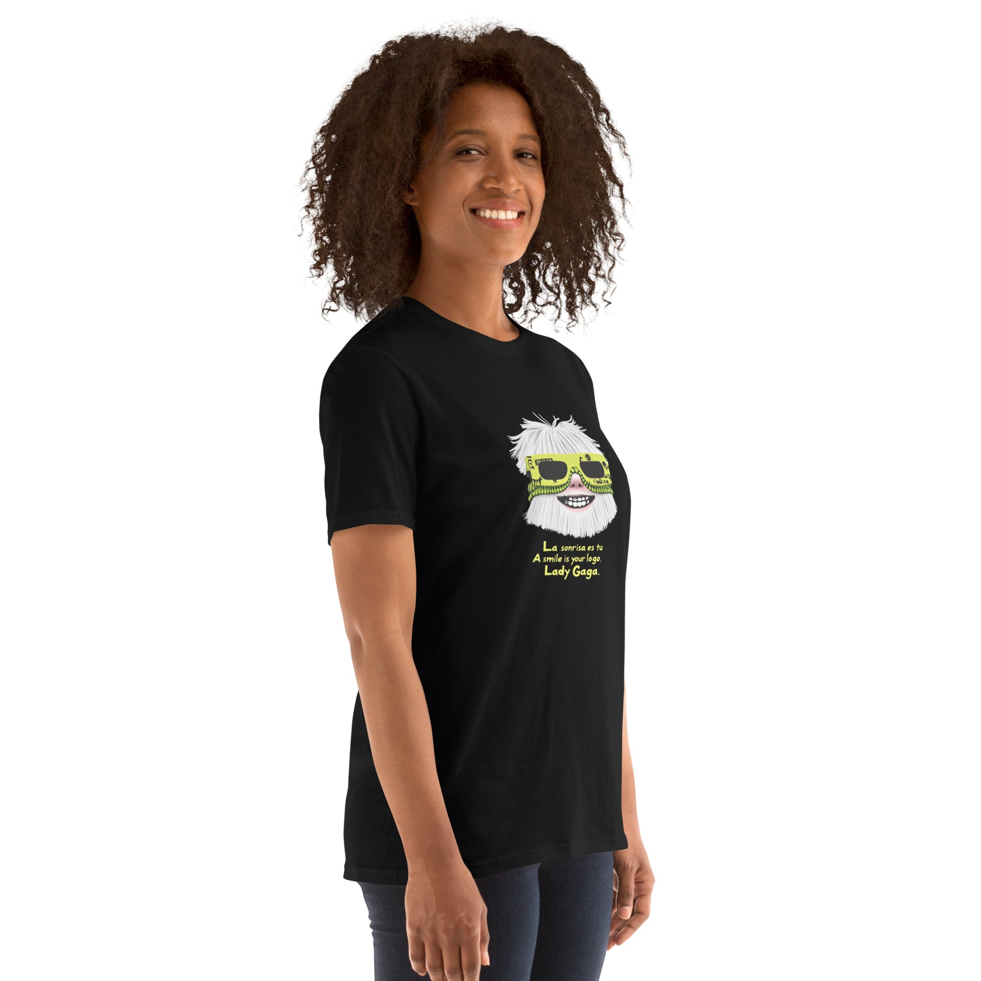 Female model wearing black T-shirt — front print: stylized face with white hair and neon sunglasses, text La sonrisa es tu / A smile is your logo. Lady Gaga.