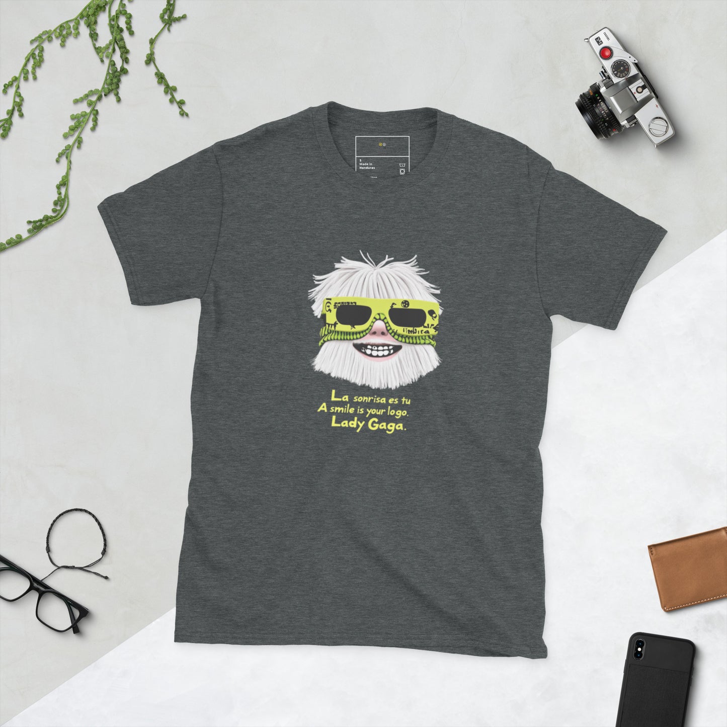 Dark grey unisex T-shirt (flat lay) — front print: stylized face with white hair and neon sunglasses, text La sonrisa es tu / A smile is your logo. Lady Gaga.