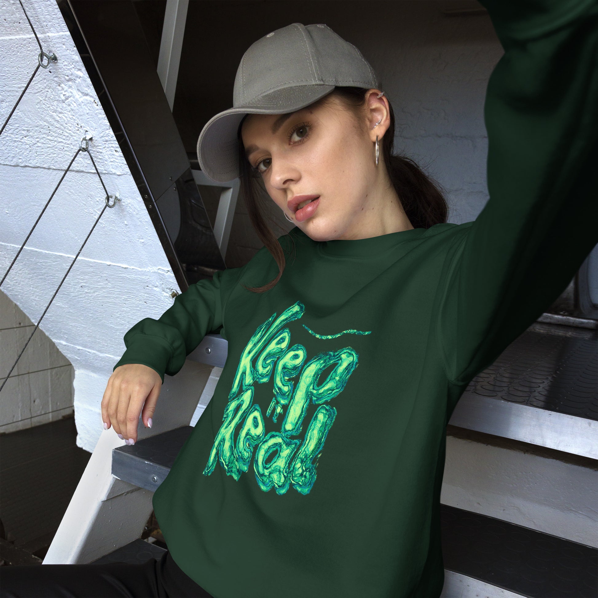 Woman in a dark green sweatshirt with “Keep Real” print, leaning casually on stairs.
